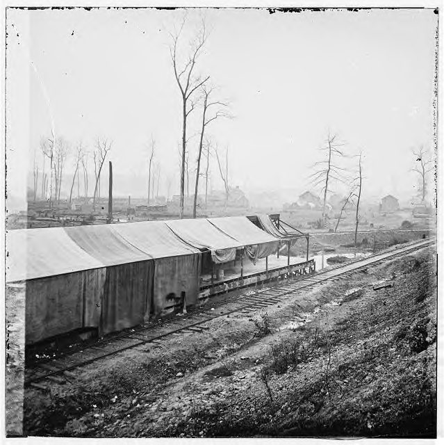 Tracks going beside a loading dock. Notice that the track is laid on ties that are laid on the bare ground. Also notice the ditching beside the track. Keeping the ditches clear and dug to the correct depth was a major job that the labor-short Confederate railroads mostly ignored during 1863 and 1864, leading to many washouts of the soil under the track and then to breaking of the line and, perhaps, derailment.